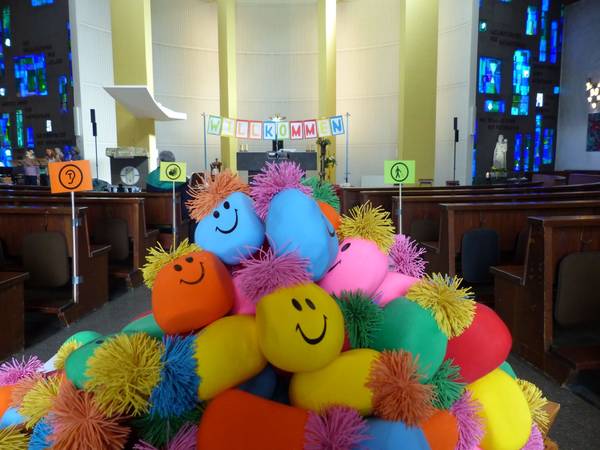 Quelle: Referat Inklusion-Generationen Es liegen bunte Knautschbälle mit Gesichtern auf einem Haufen in einer Kirche. Die Bankreihen sind für Menschen mit unterschiedlichen Behinderungen ausgeschildert.